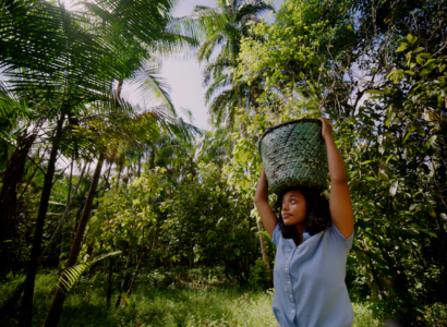 A woman carries a basket on her head through a forest
